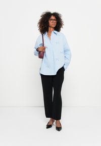 Camisa oversized azul clara com botões e detalhes bordados, combinada com calças pretas e sapatos pretos de bico fino, segurando uma pequena bolsa burgundy.