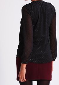 Blusa nera trasparente con maniche lunghe, motivo a punti e scollo alto. Abbinata a una gonna aderente borgogna. Tessuto testurizzato e leggero luccichio.