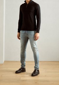 Schwarze Reißverschlussjacke, hellblaue schmal geschnittene Jeans und dunkle Mokassins mit einer Metall-Schnalle. Holzfußboden und neutraler Wandhintergrund.