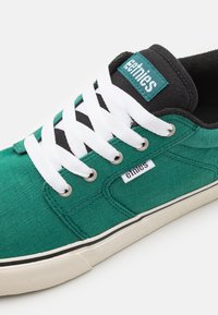 Zöld vászon sneaker fehér fűzőkkel, fekete nyelvvel, amelyen logó található, és texturált talppal, fekete kiegészítéssel.