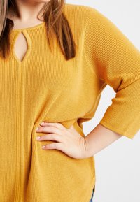 Musztardowo-żółty, luźno dziany sweter z rękawami 3/4, z dekoltem w kształcie otworu i teksturowanym materiałem.