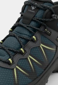 Trekkingowe buty Columbia w ciemnozielonej siateczce z czarnymi akcentami, z teksturowaną cholewką, odblaskowymi żółtymi detalami i bezpiecznym systemem sznurowania.