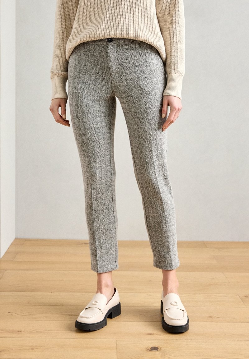 Femme portant un pantalon gris sur mesure longueur cheville, un pull beige et des mocassins crème, debout sur un sol en bois devant un mur uni.