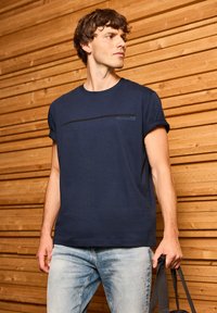 T-shirt en coton marine avec des manches retroussées, arborant un imprimé horizontal noir sur la poitrine. Le modèle tient un sac sombre, posé devant un fond en bois.