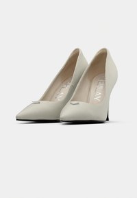 Μπεζ ψηλοτάκουνα παπούτσια με μυτερή μύτη, λείο ύφασμα και μεταλλικό διακοσμητικό στη γλώσσα. Διαθέτουν τακούνι στιλέτο και μαλακή εσωτερική επένδυση.