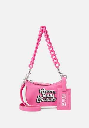 Petit sac à main rose Versace Jeans Couture avec anse chaîne épaisse, logo sur le devant et étiquette rose assortie attachée sur un fond blanc.