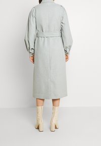 Manteau trench en mélange de laine gris avec une taille ceinturée, de longues manches bouffantes et des boutons aux poignets. Associé à des bottines crème.