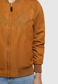 Giubbotto bomber in satin marrone con dettagli ricamati a fiori sul petto, polsini a coste e zip frontale. Indossato con jeans in denim azzurro chiaro.