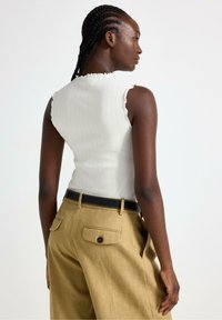 Canottiera bianca a coste con bordi scallop, abbinata a pantaloni beige a gamba larga e una cintura nera. Presenta tasche posteriori con chiusure a bottone.