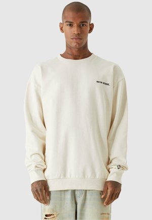 Cream-colored sweatshirt with a round neckline, dropped shoulders, and ribbed cuffs. Features small black text on the chest.