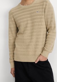 Beige gestrickter Pullover mit horizontalen strukturierten Streifen und einem runden Ausschnitt. Verfügt über gerippte Ärmelbündchen und Saum sowie ein kleines Logo auf der Brust.