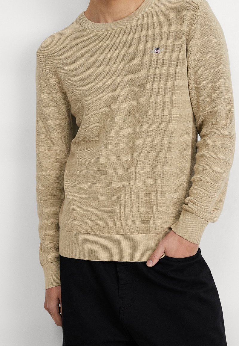 Beige gestrickter Pullover mit horizontalen strukturierten Streifen und einem runden Ausschnitt. Verfügt über gerippte Ärmelbündchen und Saum sowie ein kleines Logo auf der Brust.