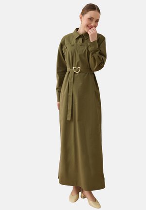 Vrouw in een lange olijfgroene jurk met ceintuur, lange mouwen en kraag, glimlachend met hand bij haar mond, draagt lichtbeige schoenen.
