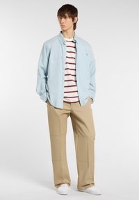 Young man stands in a casual outfit with a light blue shirt over a striped t-shirt and beige pants, against a bright background.