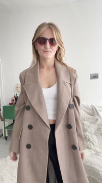 Trench coat doppiopetto in tessuto marrone chiaro con finitura strutturata, caratterizzato da grandi bottoni neri e un ampio colletto. Occhiali da sole neri indossati.