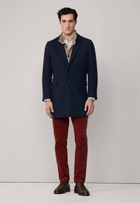 Manteau bleu marine en mélange de laine avec un design boutonné simple, un revers à pointe et deux poches avant, associé à un pantalon en velours côtelé rouge et des chaussures marron.