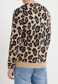 Beige tröja med ett svart leopardmönster, rund hals, ribbat ärmslut och nederkant, tillverkad av ett mjukt, texturerat stickat material.