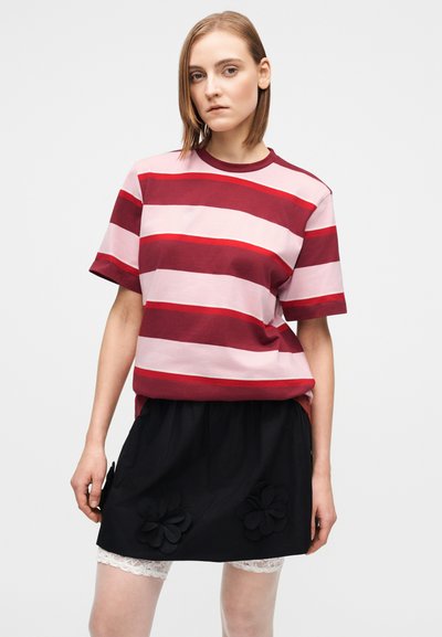 Kobieta w różowo-bordowej koszulce w paski, czarnej spódnicy z kwiatowymi aplikacjami oraz białych koronkowych szortach, stojąca na jednolitym tle.