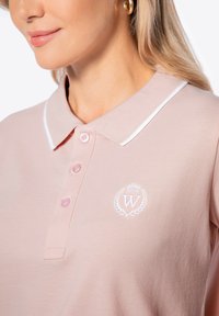 Světle růžová polo tričko s tkanou strukturou, bílým límečkem, třemi knoflíky a bílou vyšitou nášivkou na hrudi.