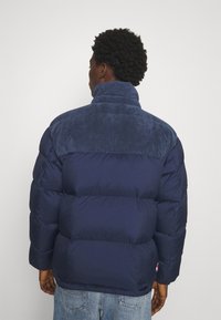 Giacca puffer blu navy con un pannello superiore in corduroy testurizzato. Presenta un collo alto e polsini elasticizzati per il calore. Etichettata con un piccolo logo.
