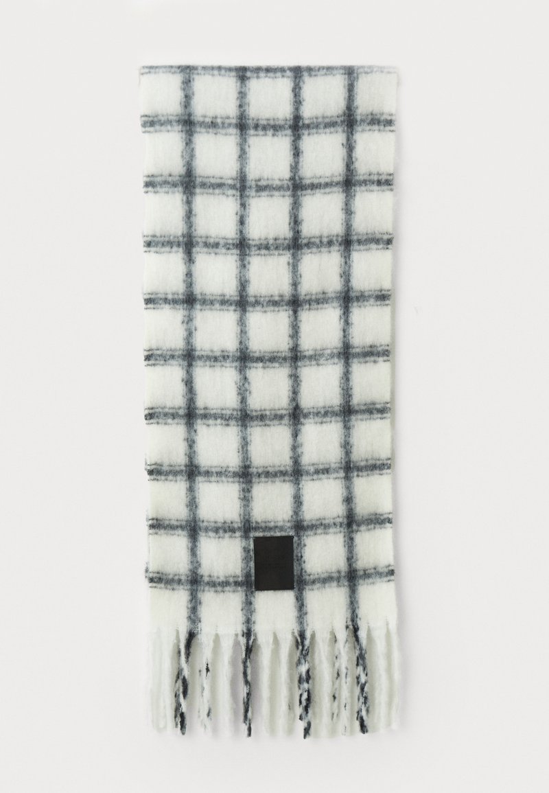 Écharpe blanche avec un motif à carreaux noirs, bords frangés et une petite étiquette noire près du bas. Fabriquée en tissu doux avec une finition texturée.