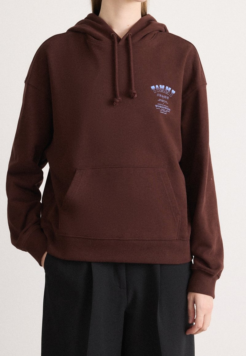 Personne portant un sweat à capuche marron avec une poche avant et un logo bleu "Tommy Jeans" sur la poitrine gauche, associé à un pantalon noir ample.
