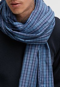 Blue plaid scarf with varying shades and fine white lines; lightweight fabric draped around the neck, contrasting with a dark textured garment.