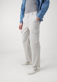 Pantalones cargo gris claro de corte recto, con bolsillos laterales y cierre de botón. Llevados con una camiseta gris y una camisa de mezclilla azul.