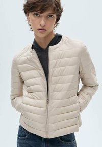Lys beige puffjakke med høj krave, horisontalt quiltet mønster og lynlås foran. Sammen med en mørk top og denimjeans.