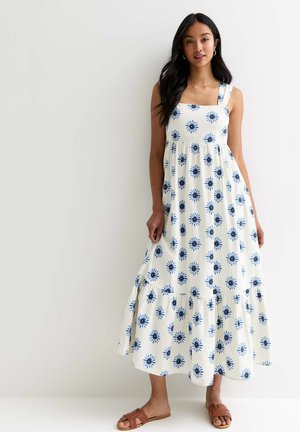 Kvinna i en vit ärmlös maxiklänning med blå blommiga mönster och bruna platta sandaler, stående mot en vit vägg.