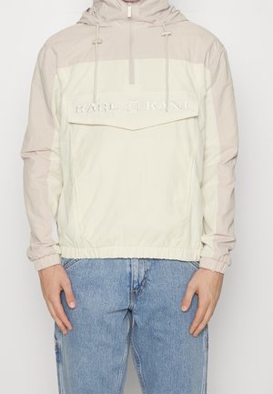 Chaqueta tipo pulóver Karl Kani en beige claro y crema con cremallera frontal y detalle de logo, usada con vaqueros azules en un modelo masculino de pie.