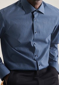 Man som bär en blåmönstrad skjorta med knappar, instoppad i svarta byxor, med händerna i fickorna, visad från halsen till höfterna.