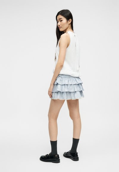 Jeune femme aux longs cheveux bruns portant un haut blanc sans manches, une jupe à volants bleu clair à étages, des chaussettes noires et des chaussures noires, vue de profil.