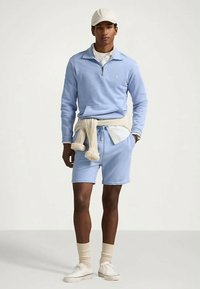 Άνδρας με ελαφρώς μπλε φούτερ με κουμπιά στο μισό ύψος και κοντά σορτς στο ίδιο χρώμα, λευκό πουκάμισο από κάτω, μπεζ πουλόβερ δεμένο γύρω από τη μέση, μπεζ καπέλο, κάλτσες και λευκά αθλητικά παπούτσια.