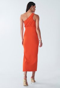 Asymmetrisches orange Kleid mit drapiertem Oberteil, tailliertem Schnitt und gerader Rockform. Kombiniert mit goldenen, schicken High Heels. Glatte Textur.
