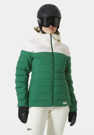 Grüne Pufferjacke mit einem weißen horizontalen Streifen, Reißverschluss und Kapuze. Kombiniert mit schwarzen Handschuhen und cremefarbenen Skihosen.