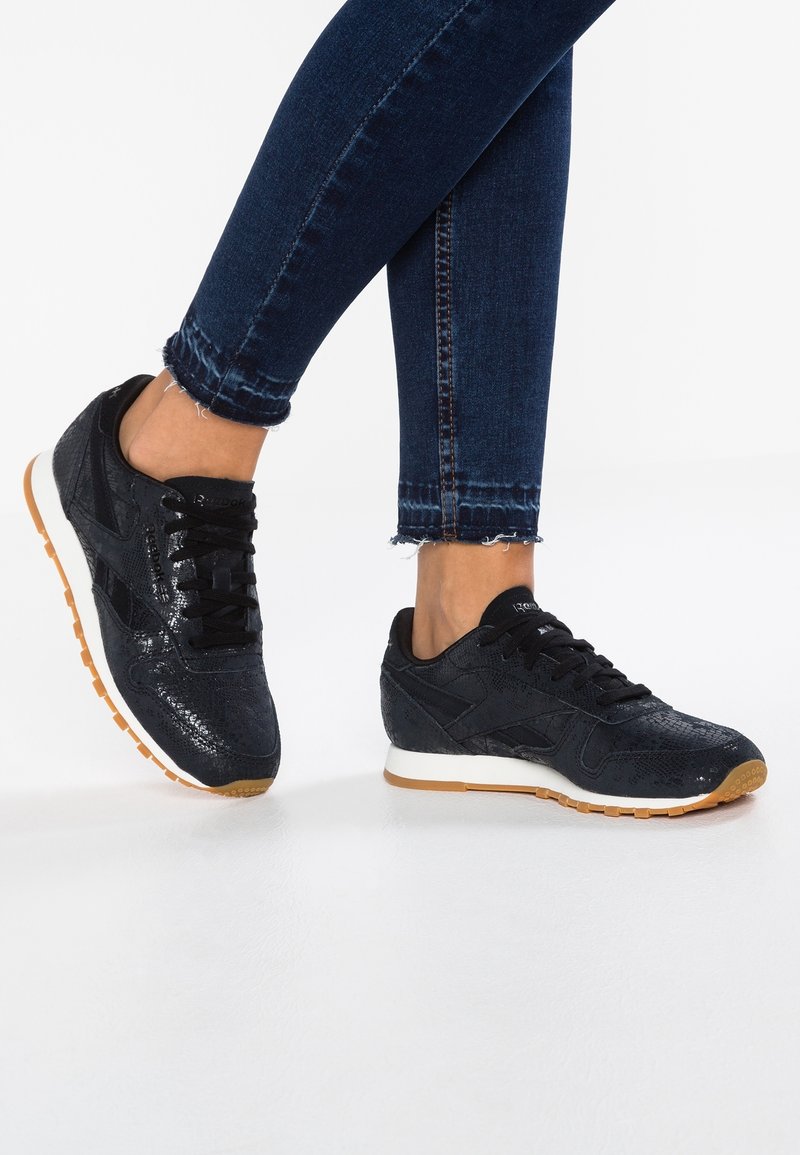 Person som bär mörkblå skinny jeans och svarta Reebok-sneakers med texturerad ovandel och gummisålar på ett vitt golv.