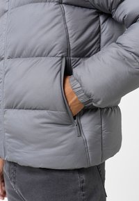 Chaqueta de plumas gris con diseño acolchado, que presenta un bolsillo lateral y puños elásticos. La textura es suave y ligeramente brillante.