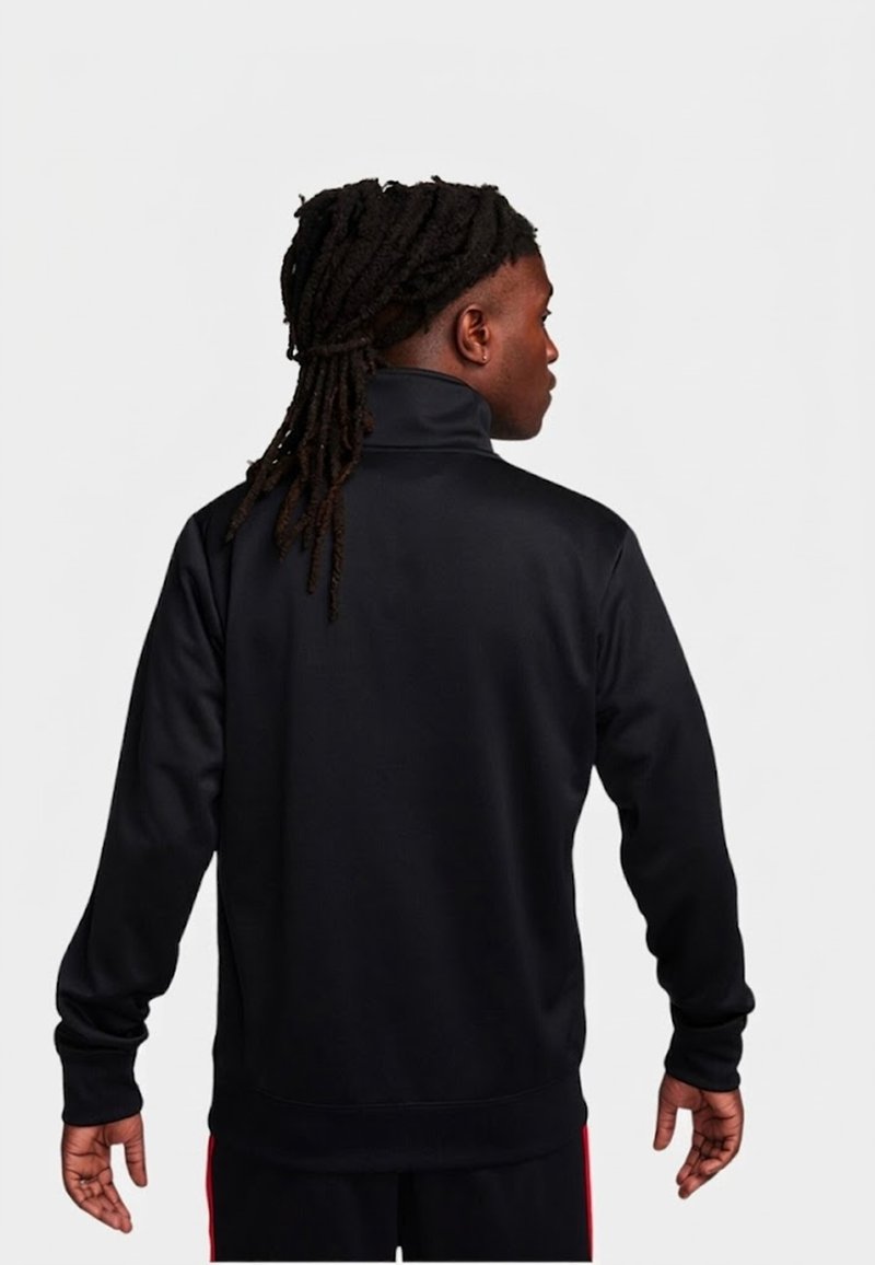 Mann mit langen Dreadlocks, der eine schwarze langärmelige Jacke trägt und mit dem Rücken zu einem schlichten weißen Hintergrund steht.
