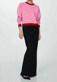 Maglione rosa con polsini e orlo rossi, abbinato a una gonna lunga nera e aderente. L'outfit presenta texture lisce e linee eleganti.