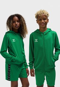 Grønne atletiske hættetrøjer med lynlås og logodetalje, kombineret med matchende grønne shorts med sort chevronmønster på siderne.