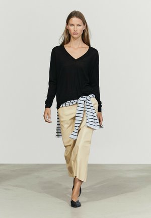 Femme portant un pull noir col en V, un pantalon beige à jambes larges, des chaussures noires, et une chemise rayée nouée autour de sa taille, marchant sur un fond uni.