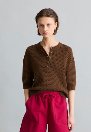 Młoda osoba z krótkimi włosami nosi brązowy, teksturowany sweter z guzikami oraz jaskrawo czerwone spodnie ze sznurkiem, stojąc na szarym tle.