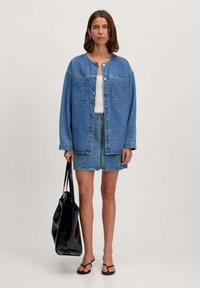 Veste en denim bleu avec une mini-jupe assortie, dotée de deux poches avant et de boutons en argent, associée à un sac fourre-tout noir et des tongs.