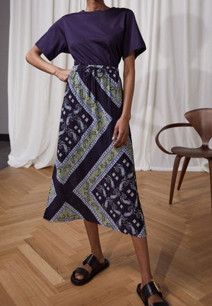 Frau trägt ein marineblaues Kurzarmtop und einen gemusterten Midirock mit grünen und weißen geometrischen Mustern, steht auf einem Holzboden.