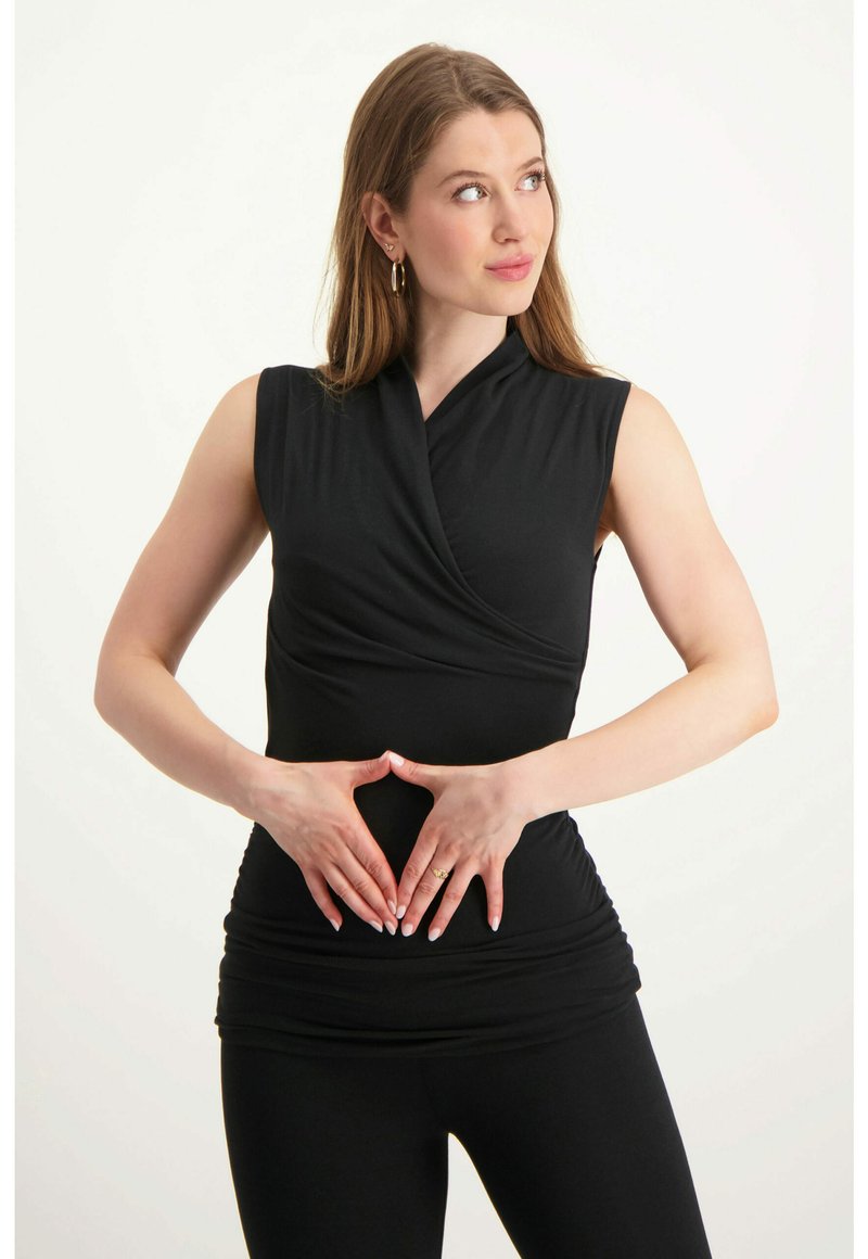Schwarzes ärmelloses Top mit gewickeltem Design und geraffter Textur an der Taille. Das Model posiert mit den Händen in Form eines Herzens auf dem Bauch.