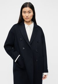 Ναυτικό διπλόκατο παλτό από μαλακό μαλλί, με μεγάλα μαύρα κουμπιά, φαρδιές πέτσες και πλαϊνές τσέπες. Λευκό πουκάμισο ορατό από κάτω.