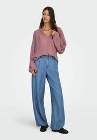 Kvinde iført en løs støvet rosa langærmet bluse med små prikker, bredbenede lyseblå bukser og sorte sko, stående med hånden nær halsen.