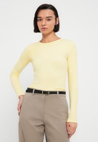 Κίτρινο ριμπ μπλουζάκι με μακριά μανίκια και στρογγυλό λαιμόκοψη, σε συνδυασμό με μπεζ παντελόνια και μαύρη ζώνη με χρυσή αγκράφα.