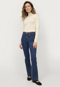 Beige coltrui, donkerblauwe flared jeans en zwarte ballet flats. Soepele textuur, slim fit bij de taille en lichte flair aan de onderkant.